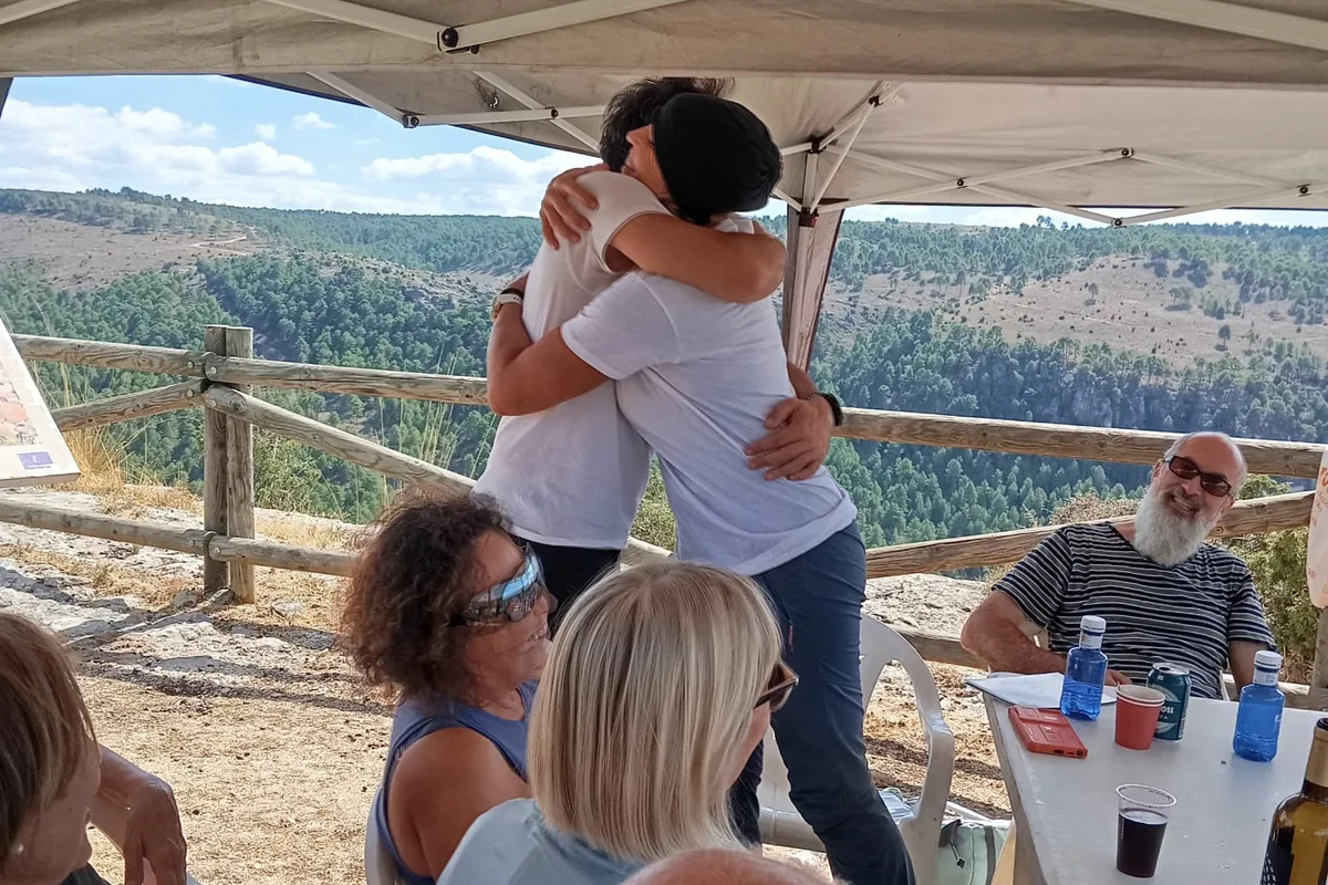 Esther y Juanma se abrazan bajo una carpa en un mirador, rodeados del grupo sentado