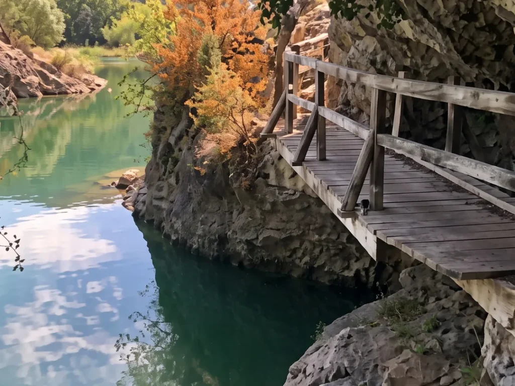 Pasarela de madera con barandilla pegada a una pared de roca caliza sobre las aguas verdes del embalse, con árboles otoñales y pinar al fondo